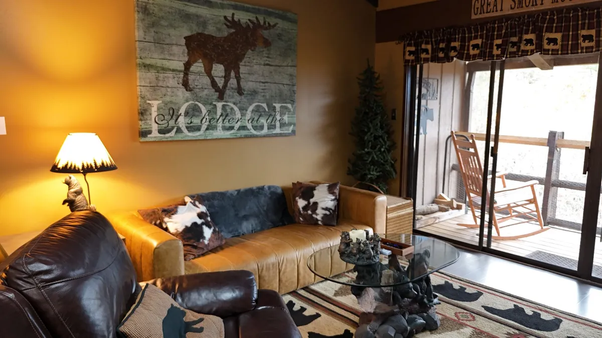 A warm and inviting living area with a brown leather sofa, glass coffee table, moose artwork, and a rocking chair. Decorative throw pillows enhance the cozy vibe, and the space feels perfect for conversation and relaxation.