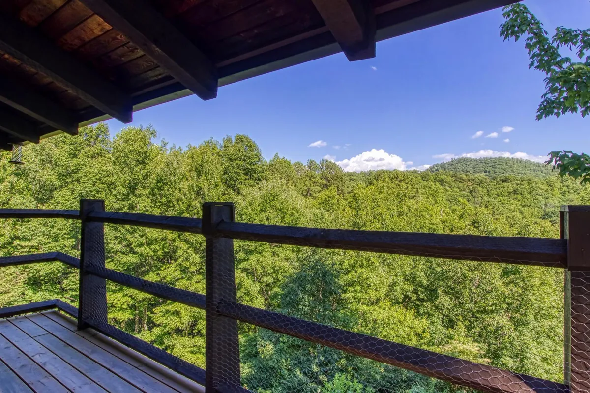 A view from a wooden balcony with a rustic railing, overlooking lush green trees and distant rolling hills. The atmosphere is serene, perfect for relaxing outdoors and observing wildlife. The deck is private and surrounded by nature, suggesting a tranquil retreat.