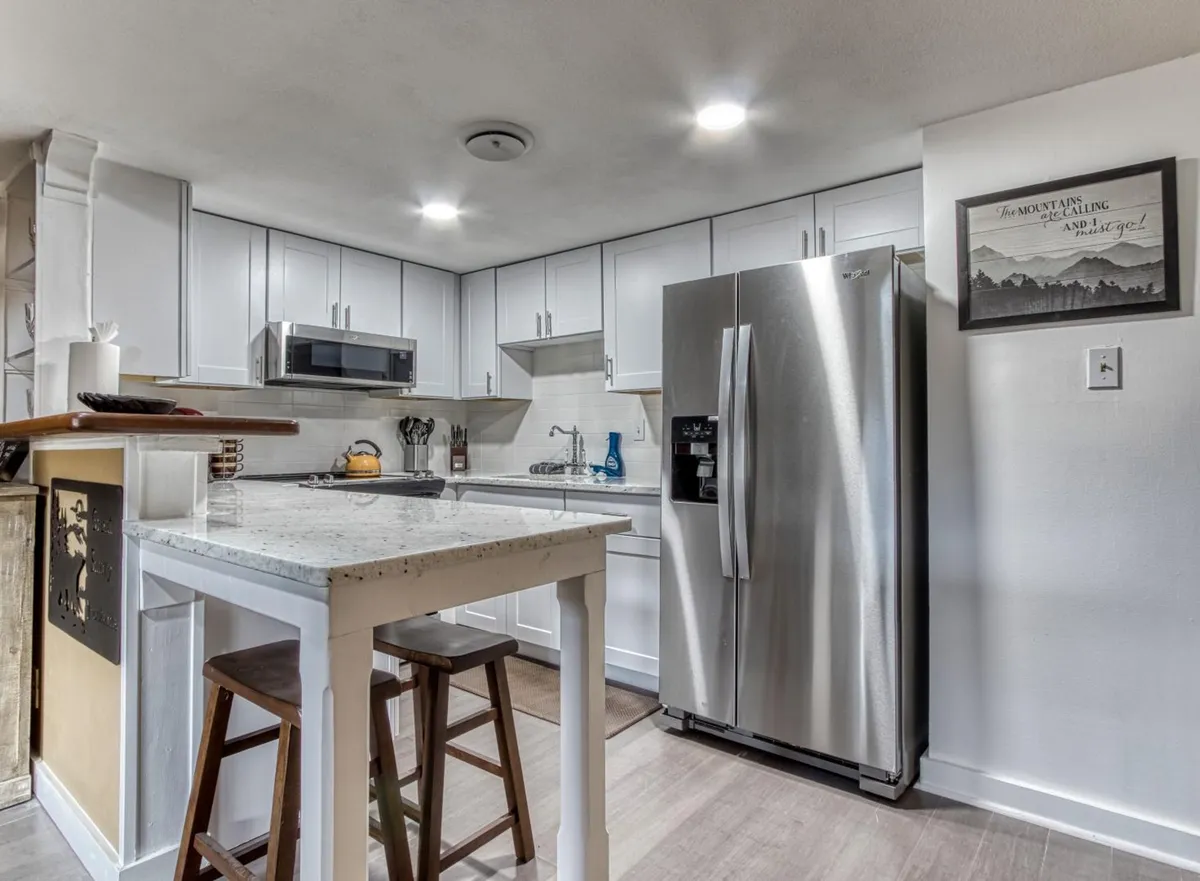 This image shows a cozy, updated kitchen featuring granite countertops, a stainless steel refrigerator, wooden bar stools, and ample cabinetry. The space is well-lit and designed for both cooking and gathering.