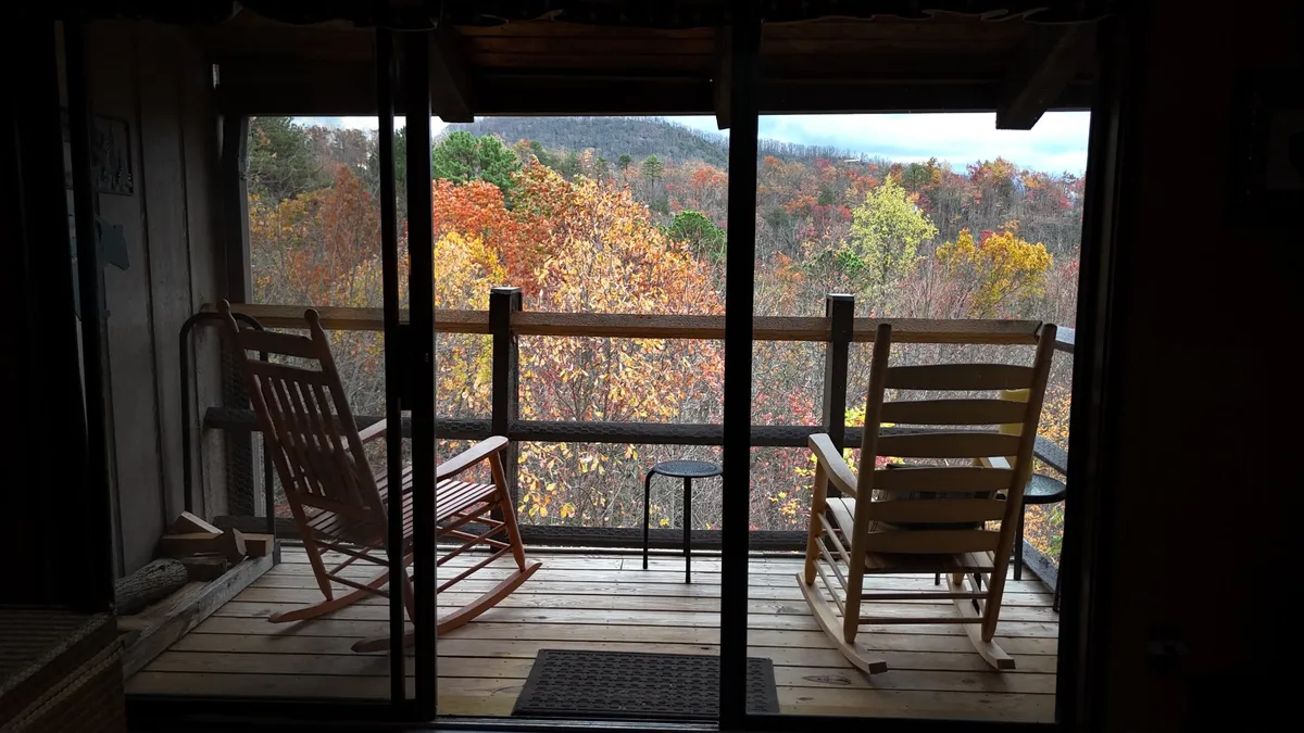 A pair of classic wooden rocking chairs are positioned on a private balcony, inviting guests to relax as they overlook a stunning expanse of autumn foliage and distant mountains. The golden hour light suggests either early morning coffee or a romantic sunset view, with the natural setting providing a peaceful, intimate atmosphere.