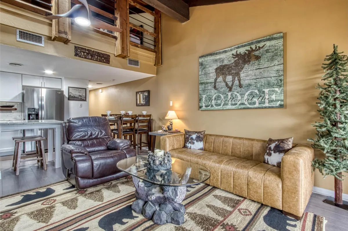 This image spotlights a leather sofa, glass coffee table, moose artwork, tree-themed décor, and a dining area. The room is arranged for both relaxing and socializing, with a warm, welcoming ambiance.