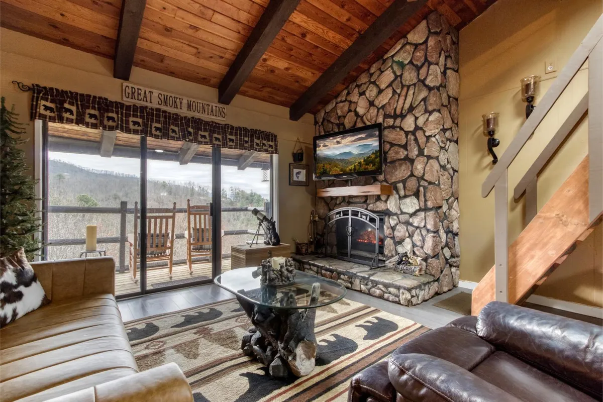 This image features a stylish interior living room with a stone fireplace, large windows that reveal beautiful mountain views, wooden beams, and rocking chairs on the balcony. The decorative rug and cozy seating create a welcoming and serene atmosphere.