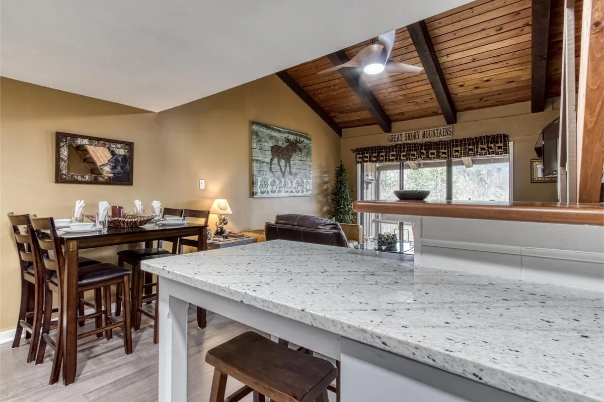 A welcoming dining area with a granite-topped table, wooden beams, moose-themed artwork, and a large window flooding the nook with natural light. The scene invites lingering over meals and enjoying the mountain surroundings.