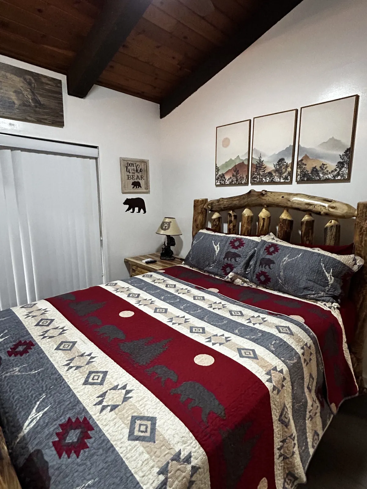 This image showcases a cozy bedroom with a log bed frame, a decorative quilt, nature-inspired artwork above the bed, and a softly glowing nightstand lamp. The room has warm wood accents, and the bedding looks inviting and plush, complemented by the ambient lighting that creates a restful atmosphere.
